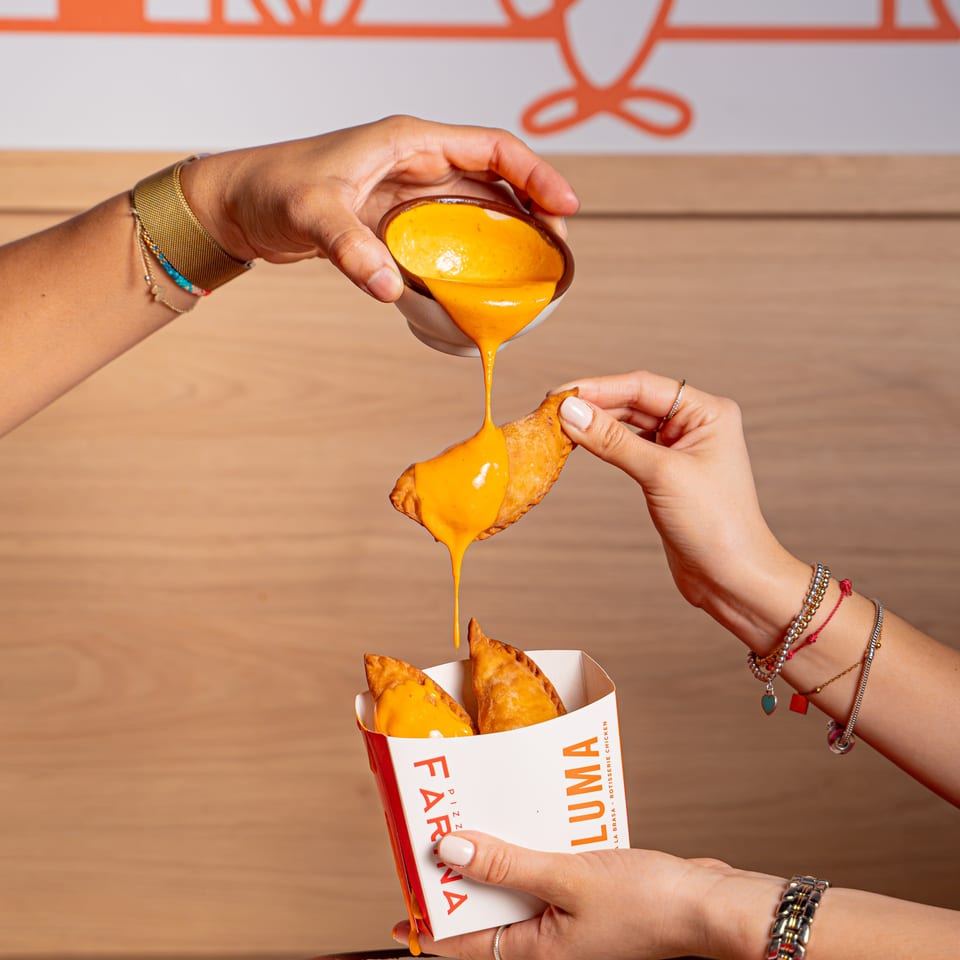 Hands drizzle creamy orange sauce over fried triangular pastries in a branded restaurant box.