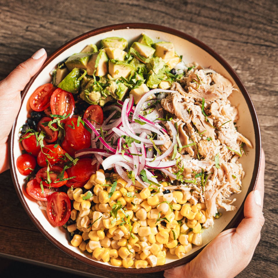 Overhead view of a colorful chicken salad with avocado, tomatoes, corn, onions, herbs, and creamy pasta held on a wooden table.