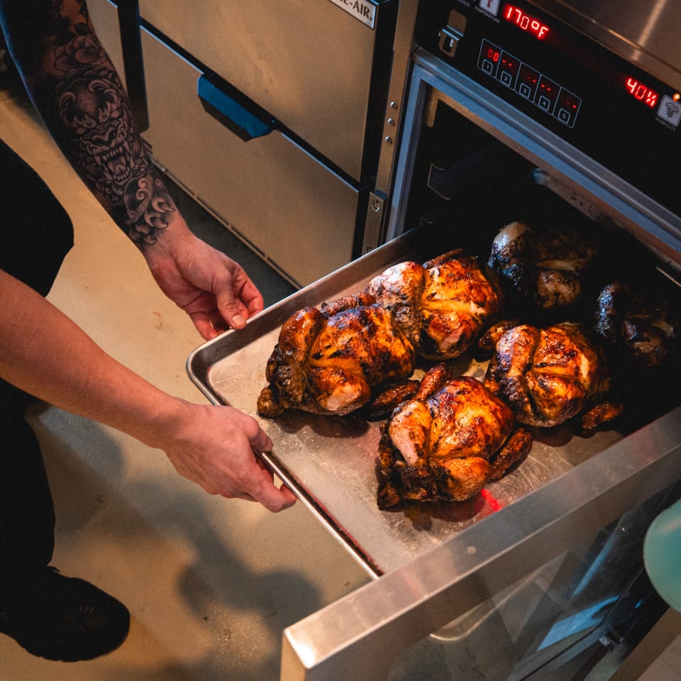 A chef with a tattooed arm places a tray of roasted whole chickens into an oven.