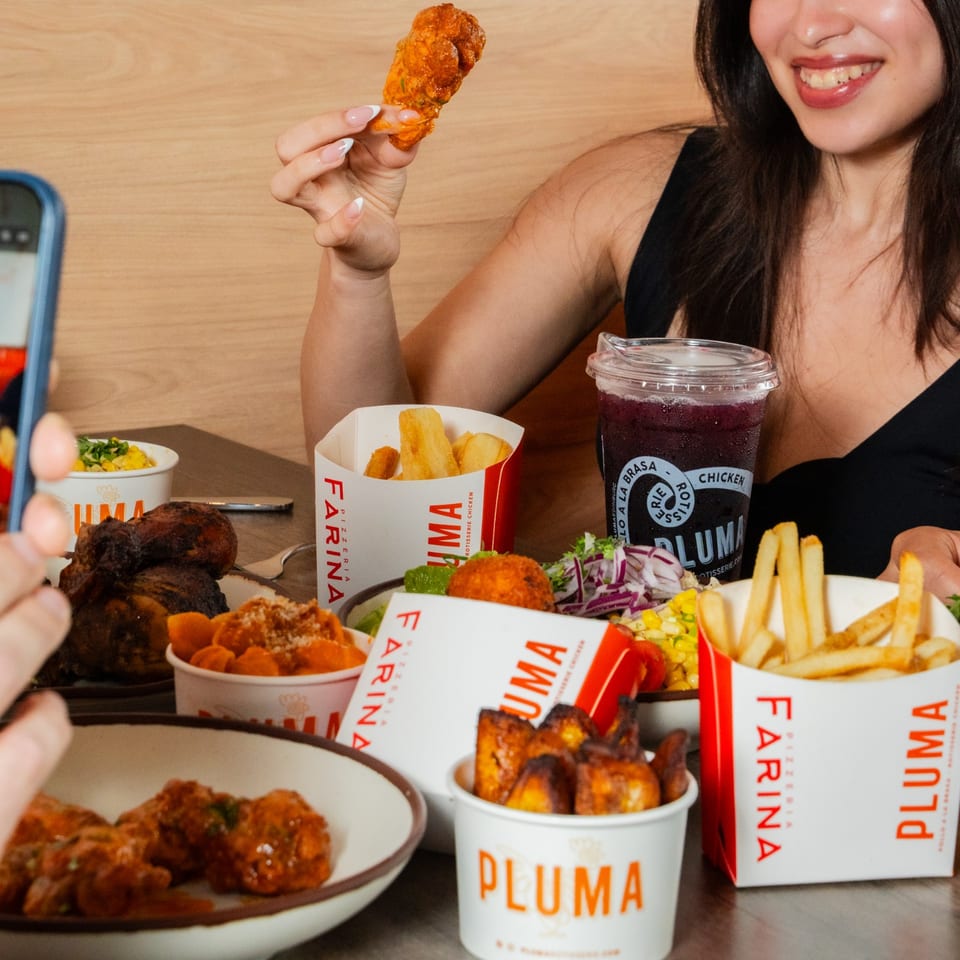 A person holds up a chicken wing, surrounded by various Pluma chicken and side dishes.