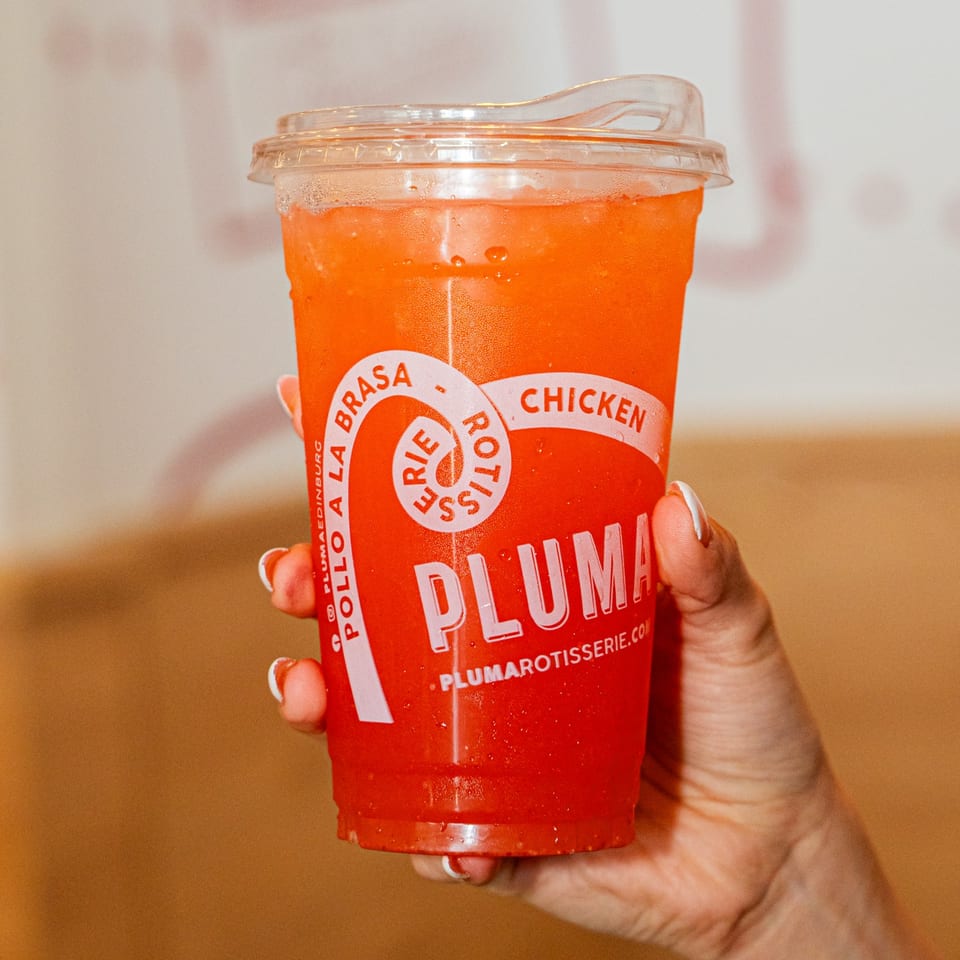 A hand holds a Pluma Rotisserie Chicken branded cup filled with a vibrant orange drink.