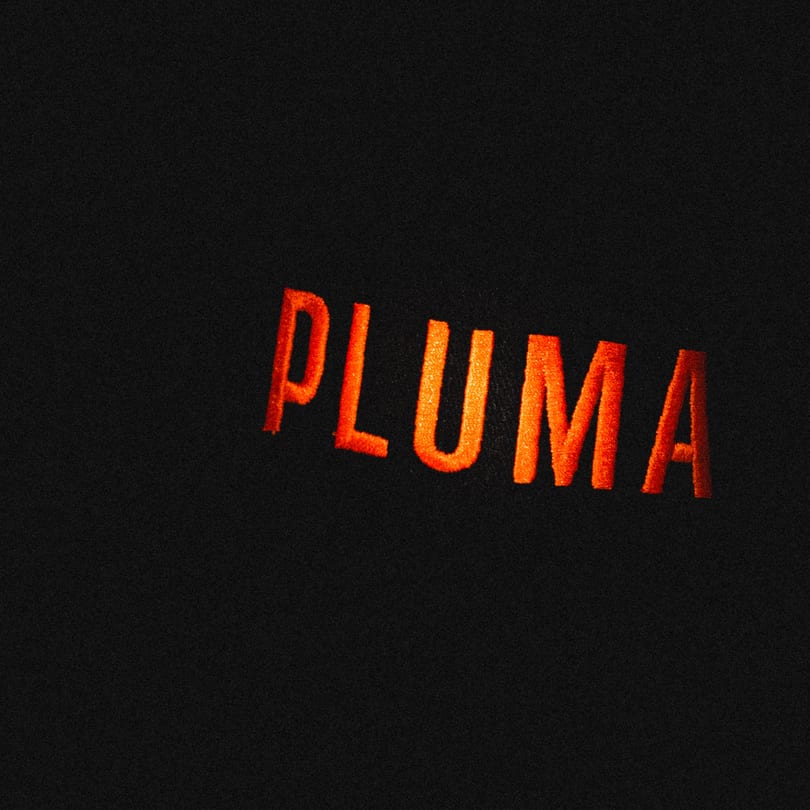 The word "PLUMA" in bright orange, textured letters against a dark, grainy background.