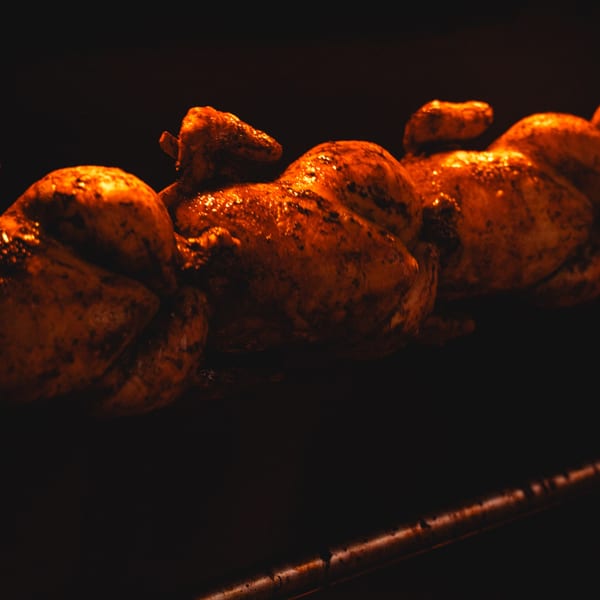 Three rotisserie chickens, golden brown and glistening, cooking on a spit in a dark oven.