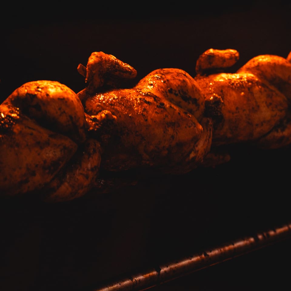 Three rotisserie chickens, golden brown and glistening, cooking on a spit in a dark oven.